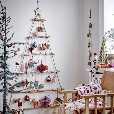 Arbre de Noël en bois et corde sur lequelle sont accrochées de décorations de Noël, le tout à côté d'un sapin de Noël et d'une chaise en bois sur laquelle est posé en désordre un plaid.