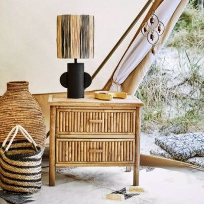 Chevet 2 tiroirs en bambou dans une tente ouverte sur lequel se trouve une lampe avec un abat-jour en raphia noir et naturel avec un socle noir avec deux oreilles