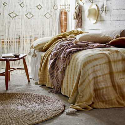 Chambre avec un lit désordonné sur lequel il y a des coussins et un plaid à côté duquel on trouve une table de chevet en bois et un tapis en herbier