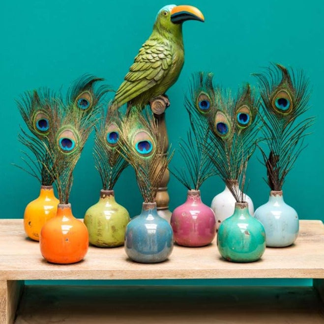 Assortiment de vases colorés dans lequel sont posées des plumes de paon, le tout sur une table en bois clair.